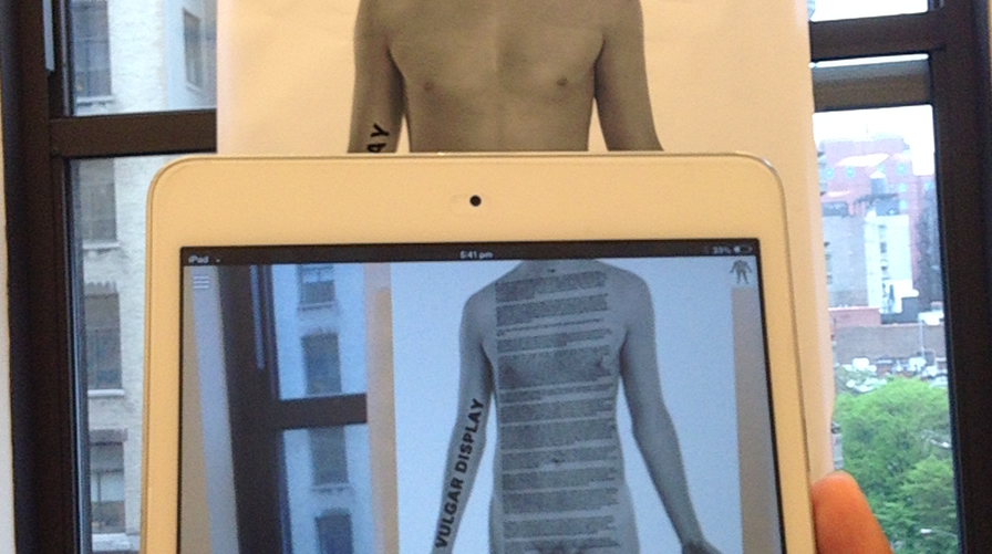 The image shows a tablet being held up in front of a photograph of a man's torso. The augmented reality experience on the tablet adds a text overlay to the torso, so the viewer can read the text as if it were printed on the torso.