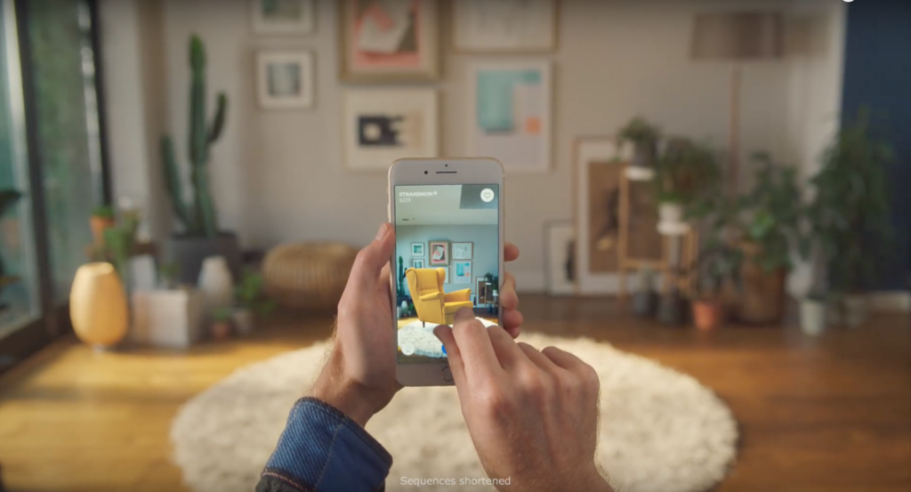 Image shows a person's hands holding a mobile phone. The person is in a living space, and the image on the phone is of that living space with an Ikea wing chair added to the scene.