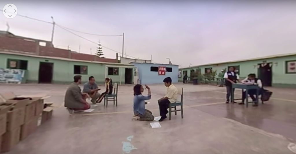 Image shows an outdoor, open plaza with one-storey buildings in the background. People are on the plaza clustered in groups of people kneeling facing people sitting in chairs.